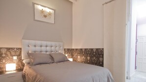 A bedroom with a tufted headboard, a bed with a grey duvet, two bedside tables with lamps, and a patterned wallpaper on the lower half of the walls. - Imperial Deluxe Plaza Mayor (Madrid)