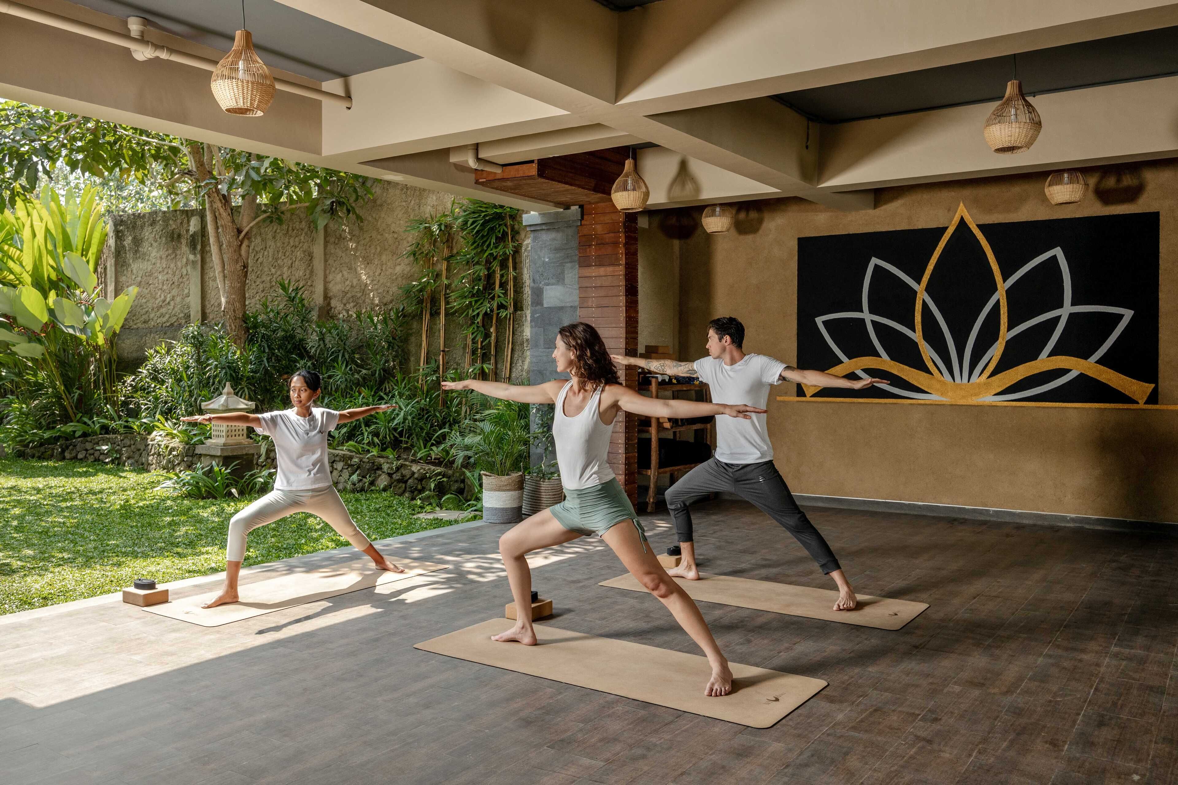 A yoga studio with three people practicing on mats, surrounded by greenery and a large wall mural.
