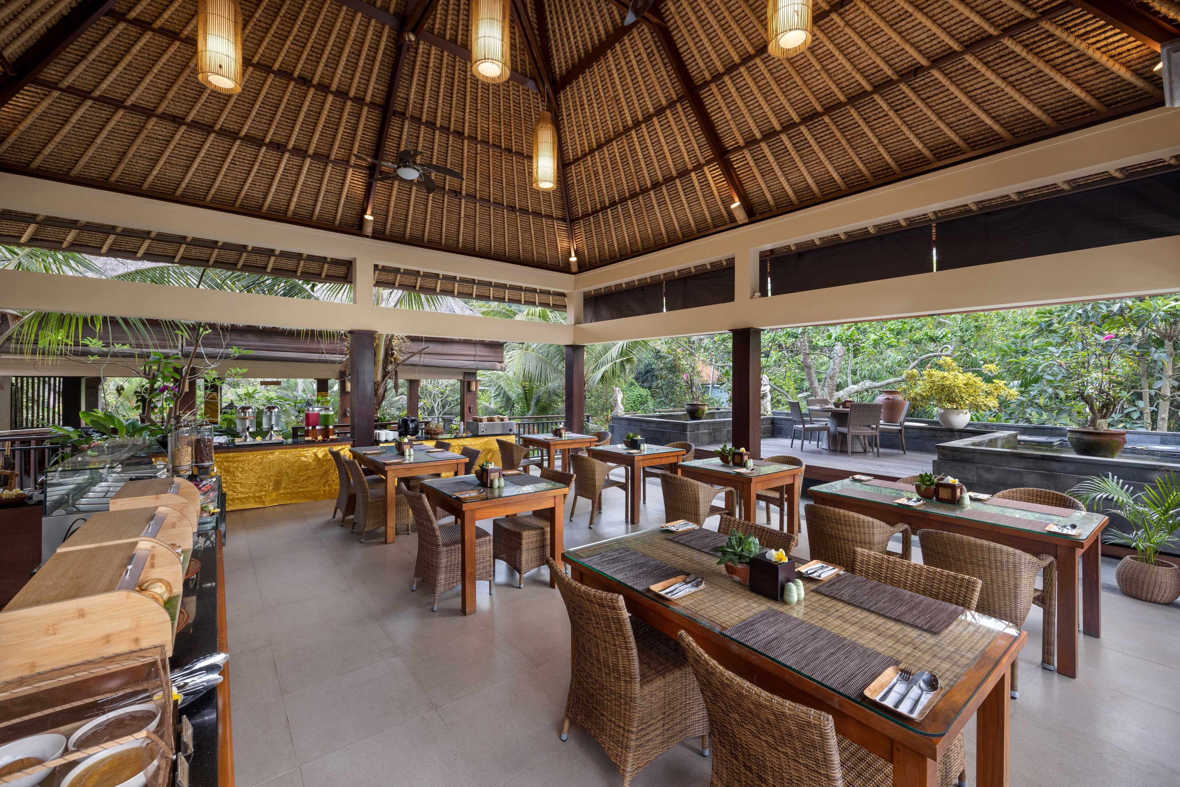 An outdoor dining area with wooden tables and wicker chairs, surrounded by potted plants and a thatched roof.