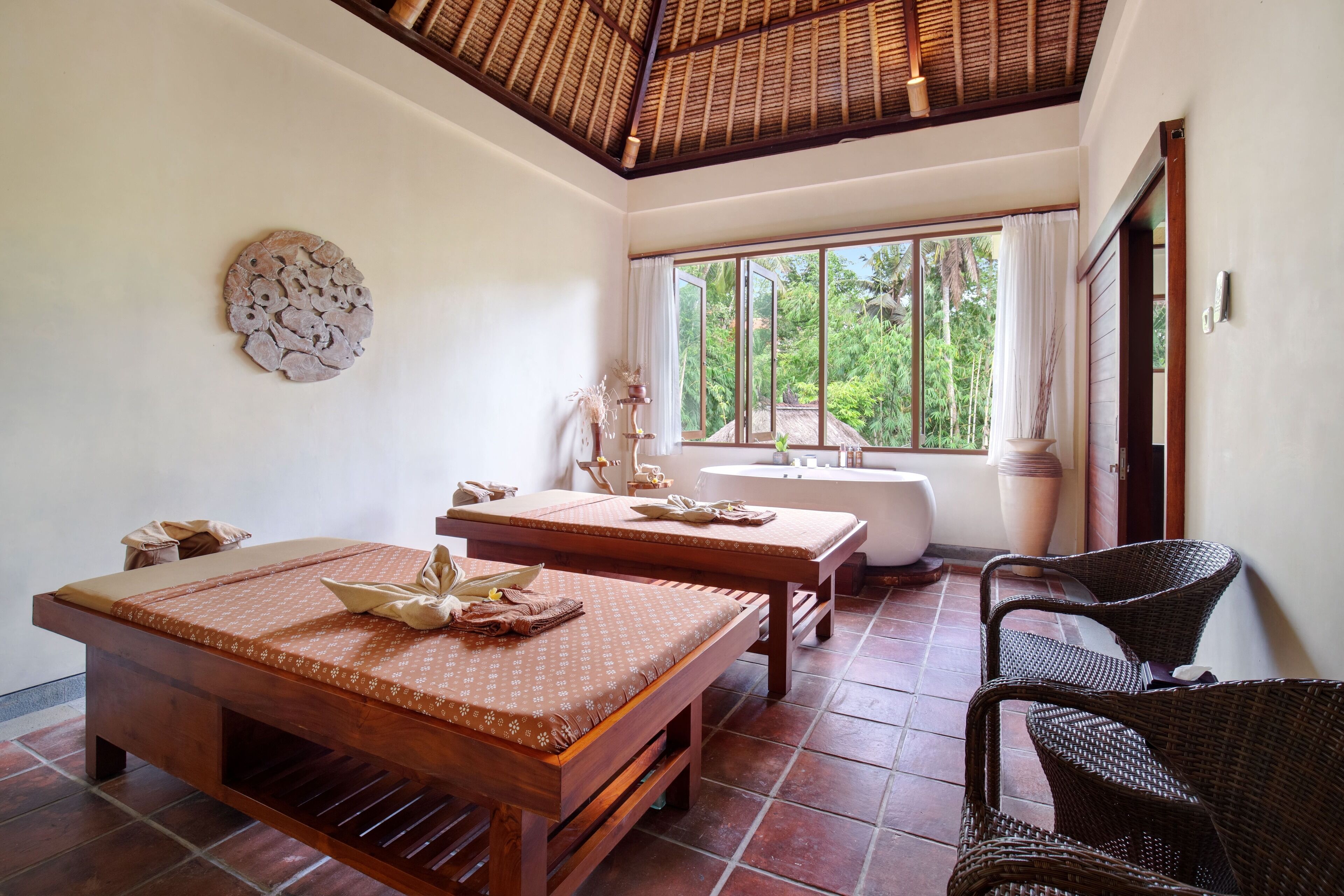 A room with a wooden bed, a wooden table, wicker chairs, a large window, and a ceiling with a woven design.