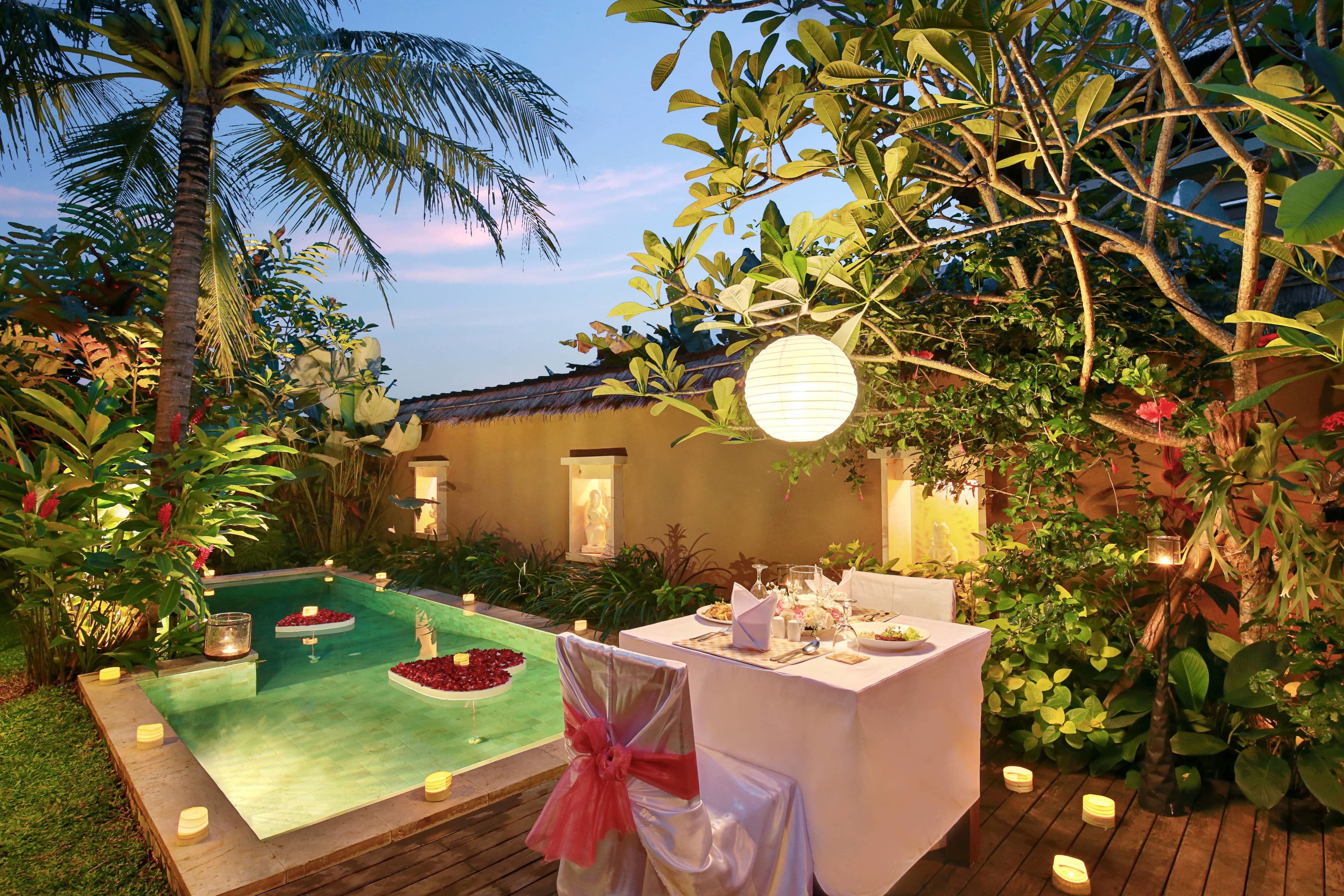 An outdoor dining area with a pool, surrounded by lush greenery and palm trees.