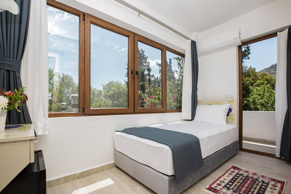 Ein Schlafzimmer mit Bett, großen Fenstern, Blick ins Grüne und einem kleinen Tisch mit einer Vase.