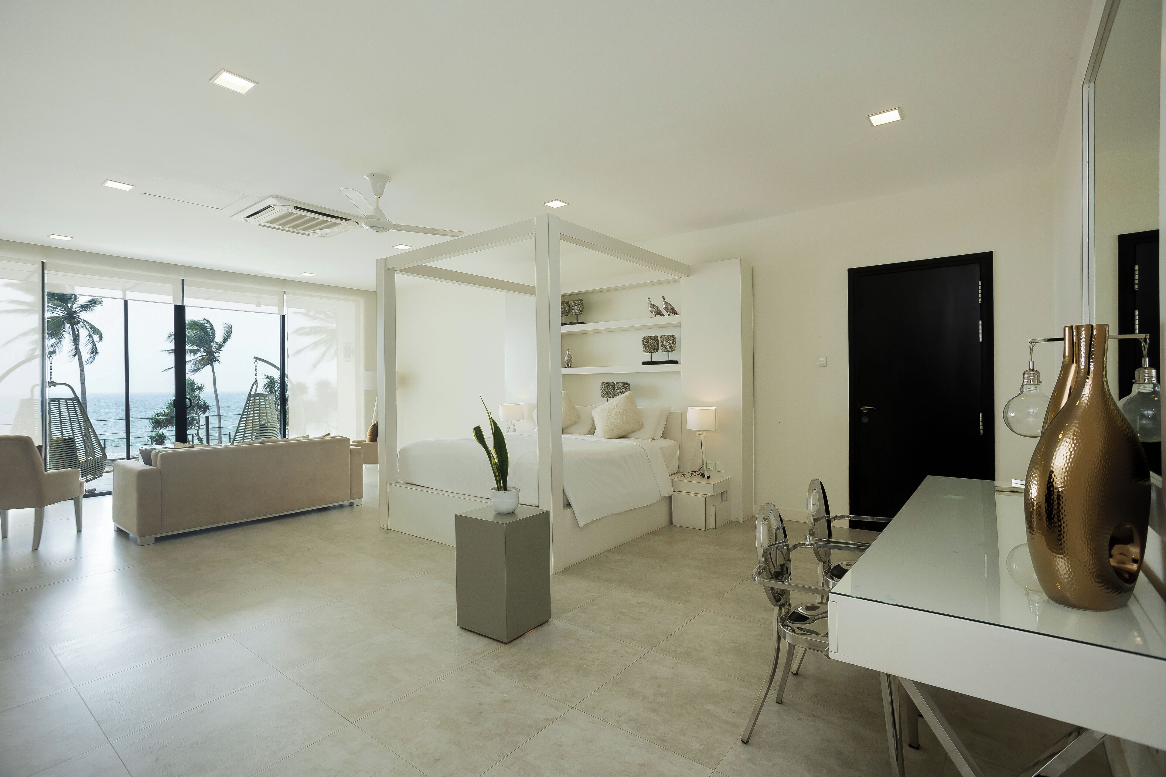 A modern hotel room with a large bed, a sitting area, a dining table, and a view of palm trees.