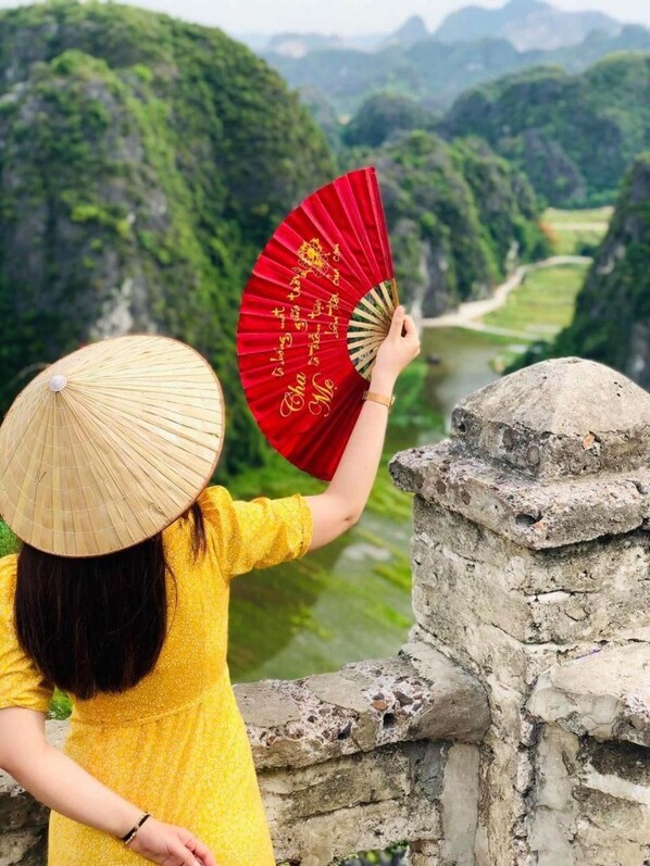 Una persona sostiene un abanico rojo con diseños dorados, de pie sobre una estructura de piedra con vistas a un valle verde.