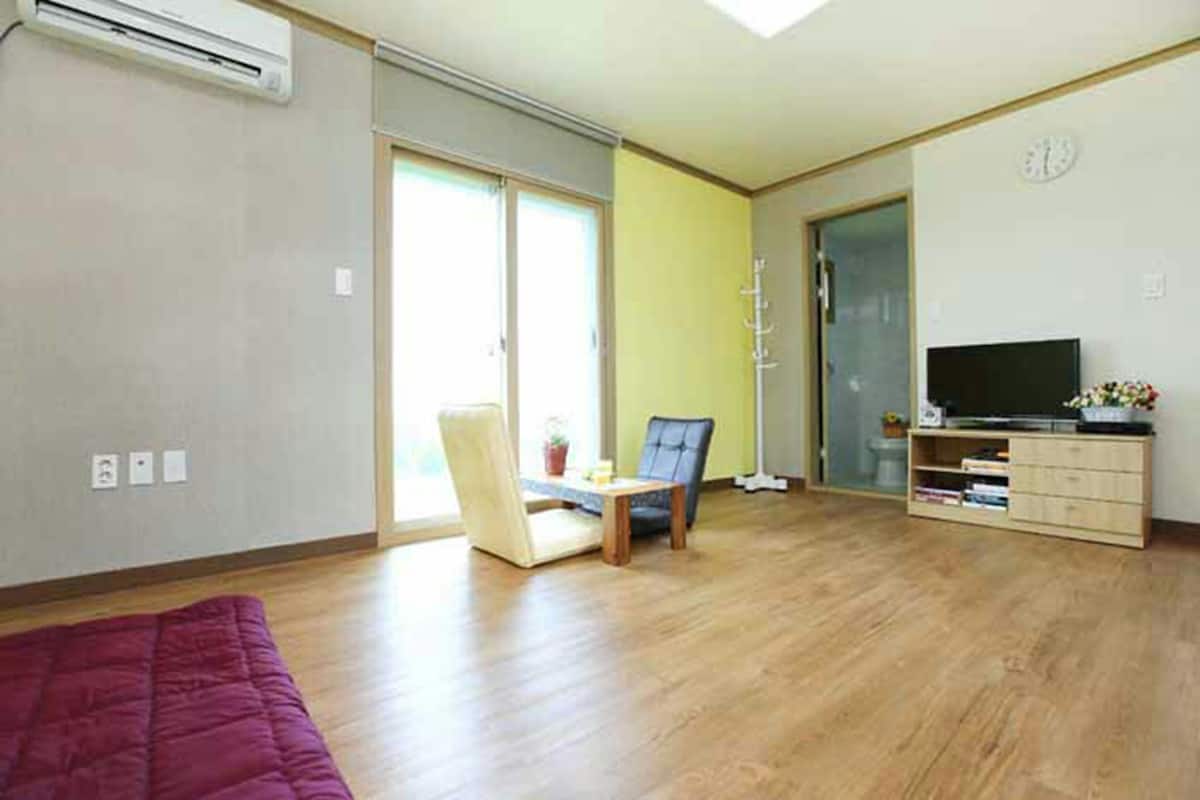 A spacious living room with a large window, a flat-screen TV, a wooden cabinet, and a purple sofa.