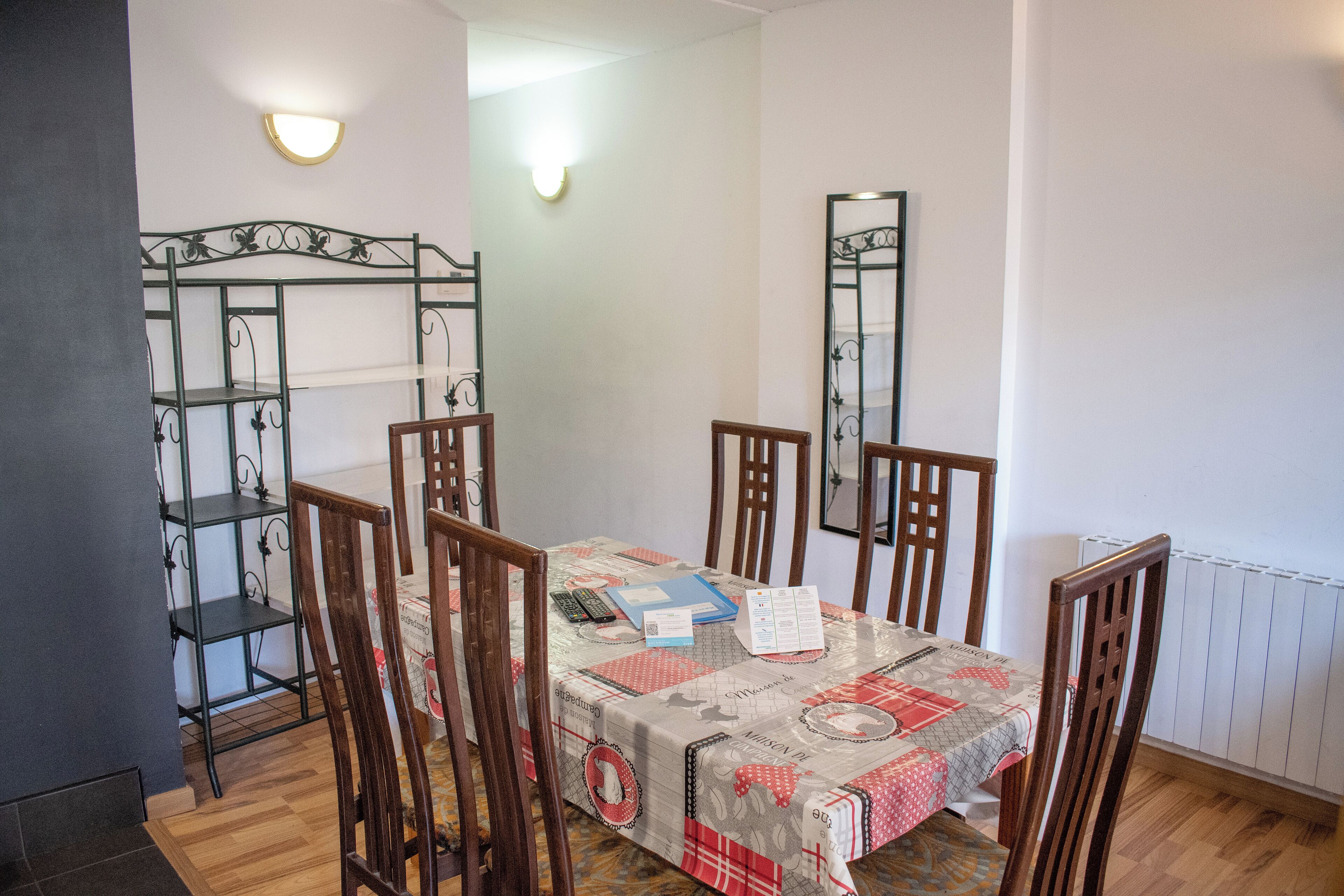 A dining area with a table set for four, surrounded by wooden chairs. A metal shelf unit is to the left, and a mirror hangs on the wall.