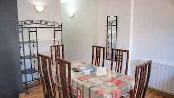 A dining area with a table set for four, surrounded by wooden chairs. A metal shelf unit is to the left, and a mirror hangs on the wall.