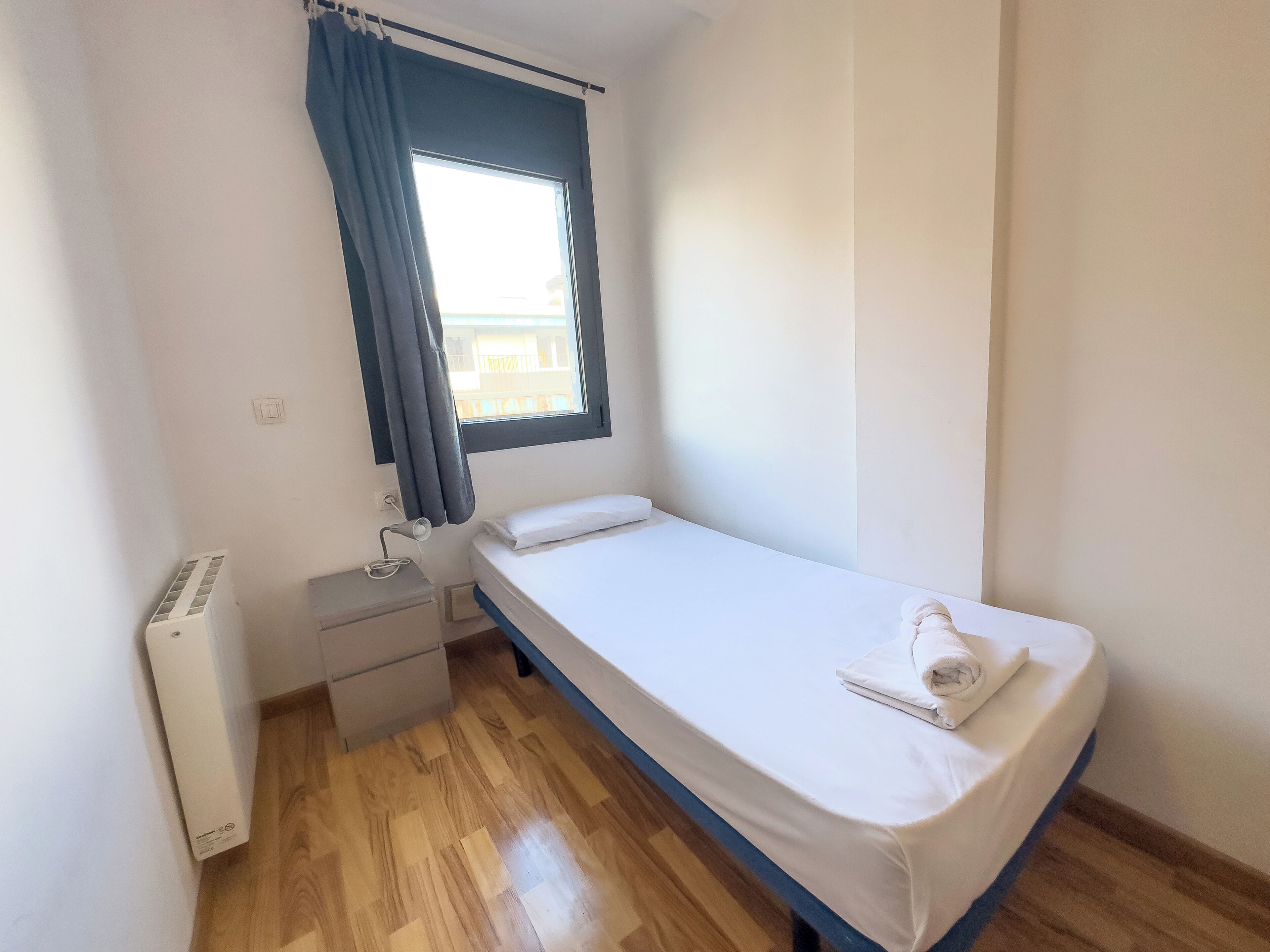 A small, clean room with a single bed, a bedside table, a heater, and a window with curtains.