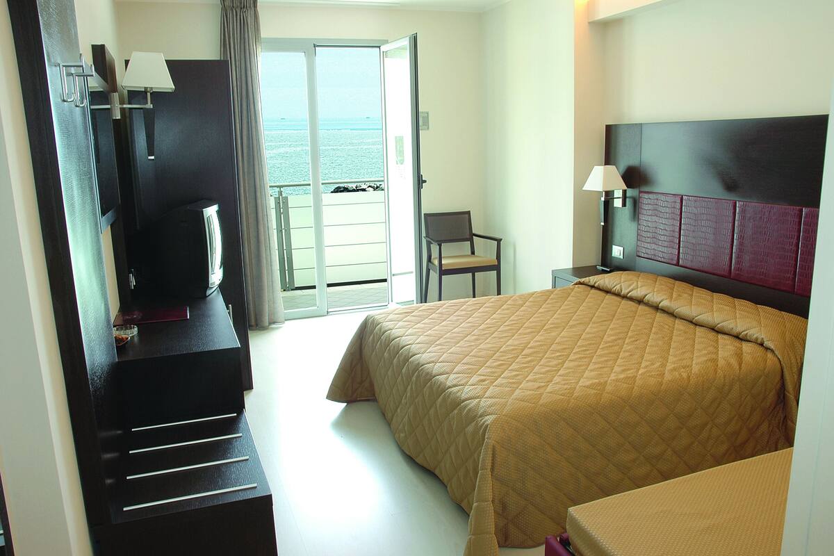 Een hotelkamer met een bed, een bureau met een televisie, een stoel en uitzicht op zee door een schuifdeur.