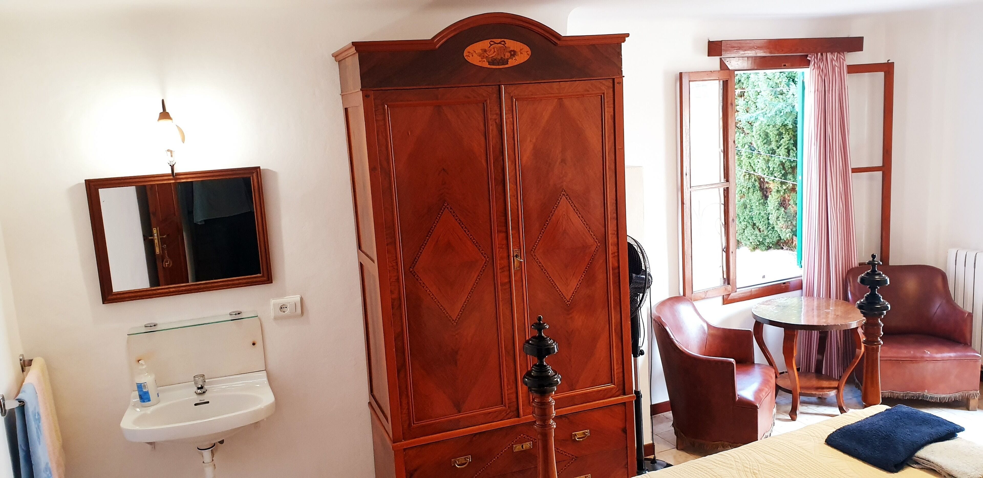 A room with a wooden wardrobe, a mirror, a sink, a chair, a table, and a window with curtains.