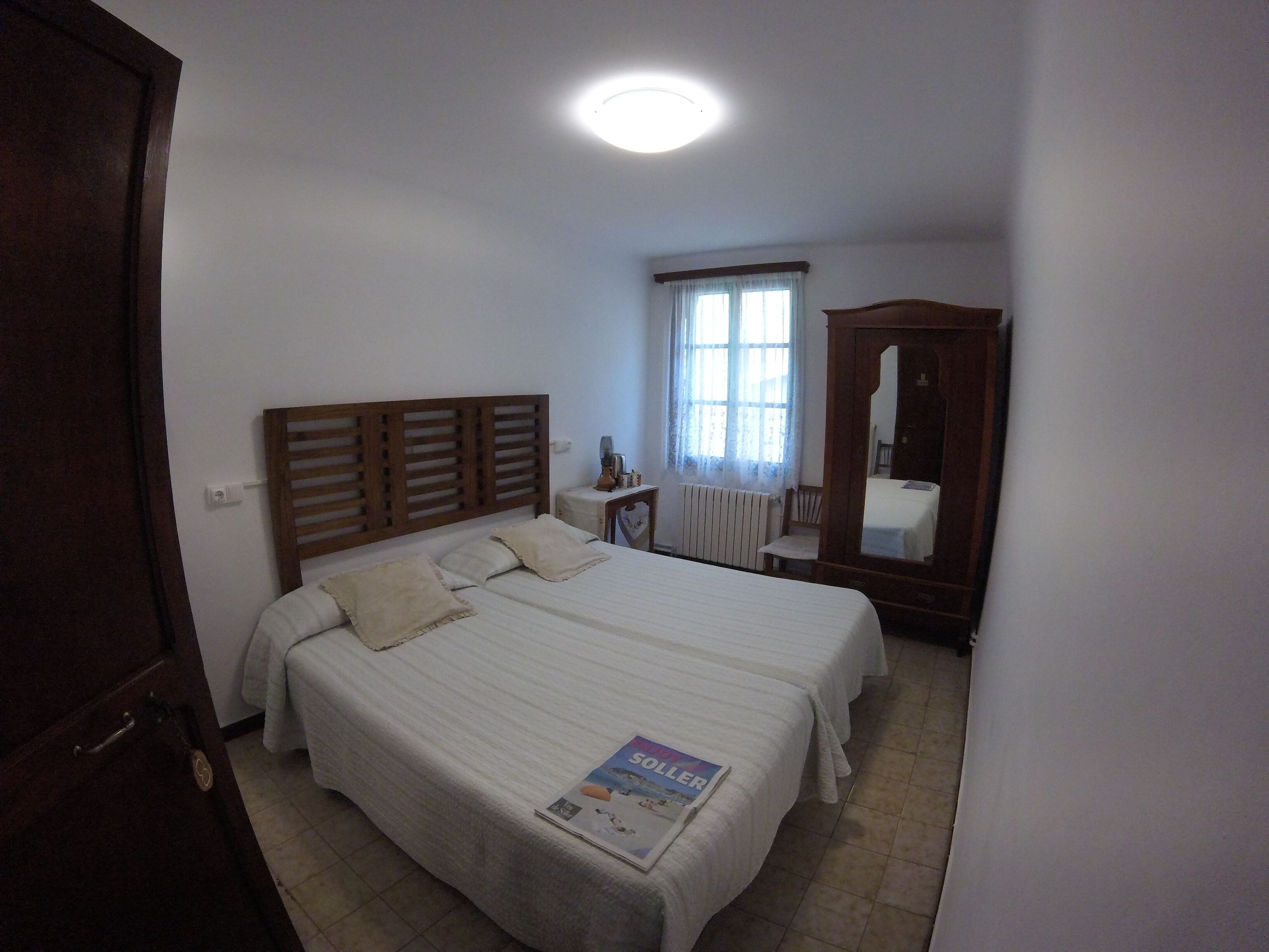 A bedroom with a large bed, a wooden headboard, a bedside table with a lamp, a window with curtains, and a mirror.