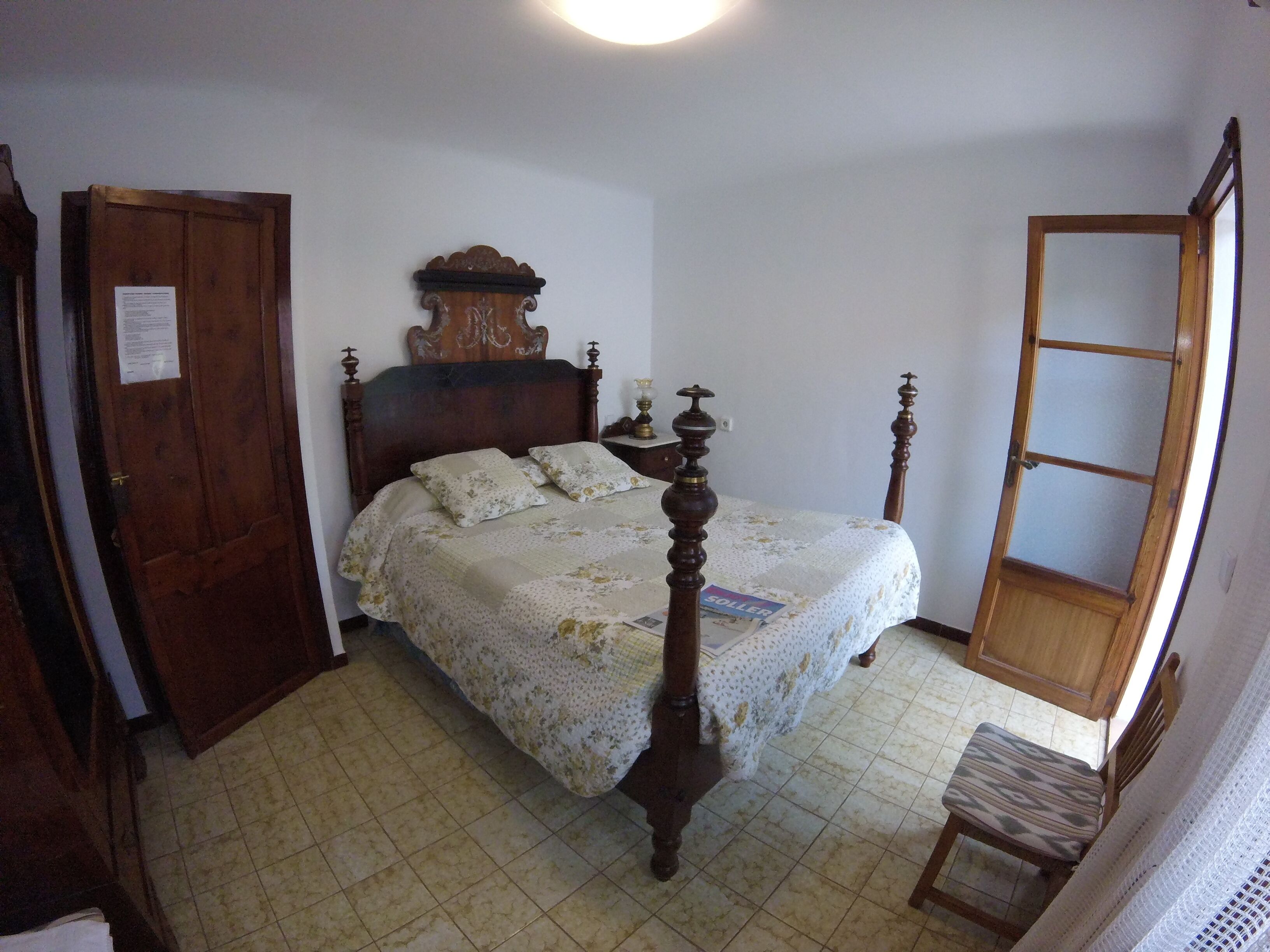 Een slaapkamer met een hemelbed, een houten deur, een klein tafeltje met een lamp en een stoel.