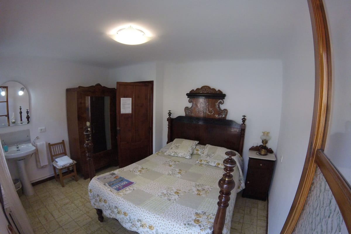 Quarto com cama de madeira, mesa de cabeceira, espelho, guarda-roupa de madeira e banheiro com pia e cadeira.