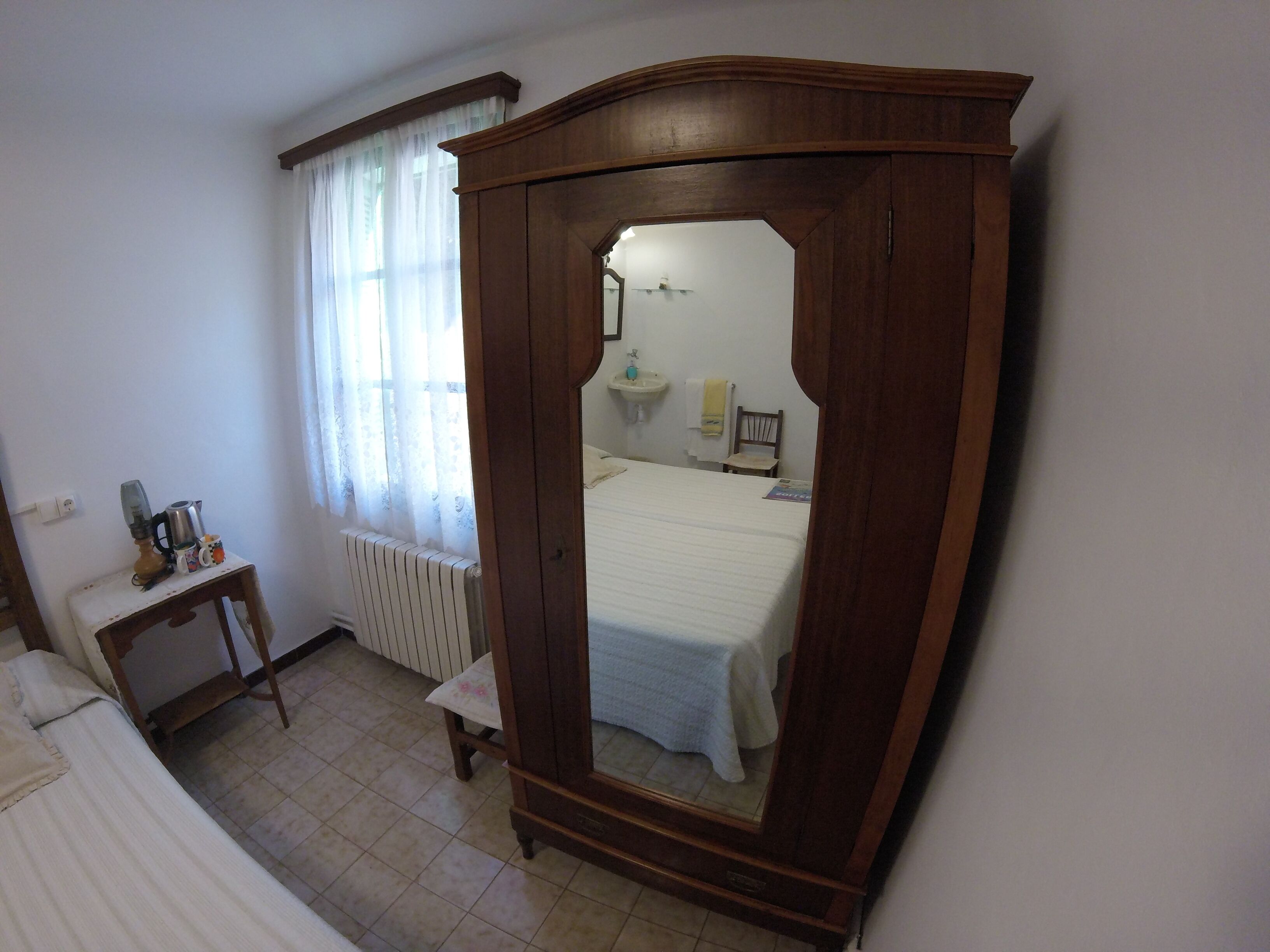 Quarto com guarda-roupa de madeira, uma pequena mesa de cabeceira, radiador, janela com cortinas finas e cama com edredom branco.