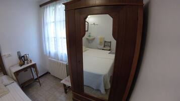 Quarto com guarda-roupa de madeira, uma pequena mesa de cabeceira, radiador, janela com cortinas finas e cama com edredom branco.