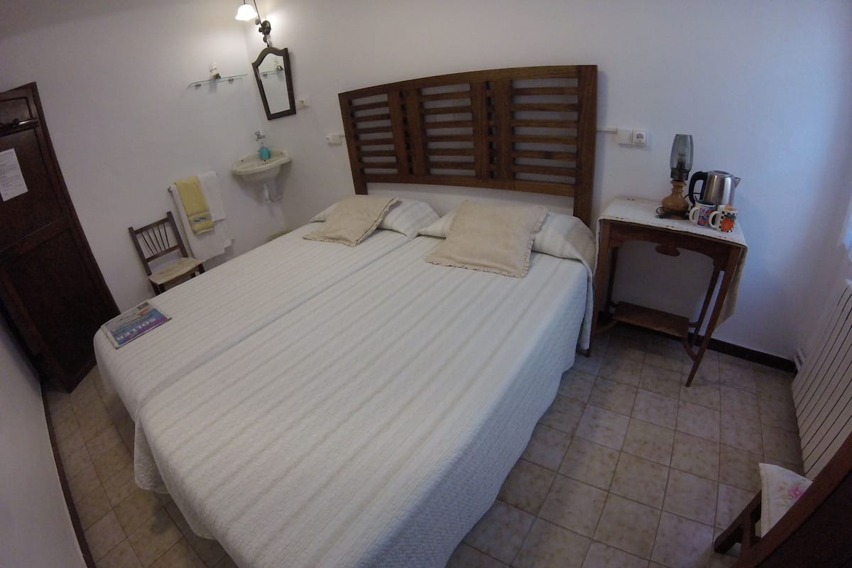 Cama bem arrumada com cabeceira de madeira, mesa de cabeceira com chaleira e prateleira pequena com espelho e uma garrafa.