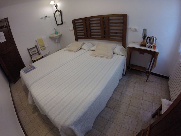 Una cama bien tendida con cabecera de madera, una mesita de noche con tetera, y un pequeño estante con espejo y botella.