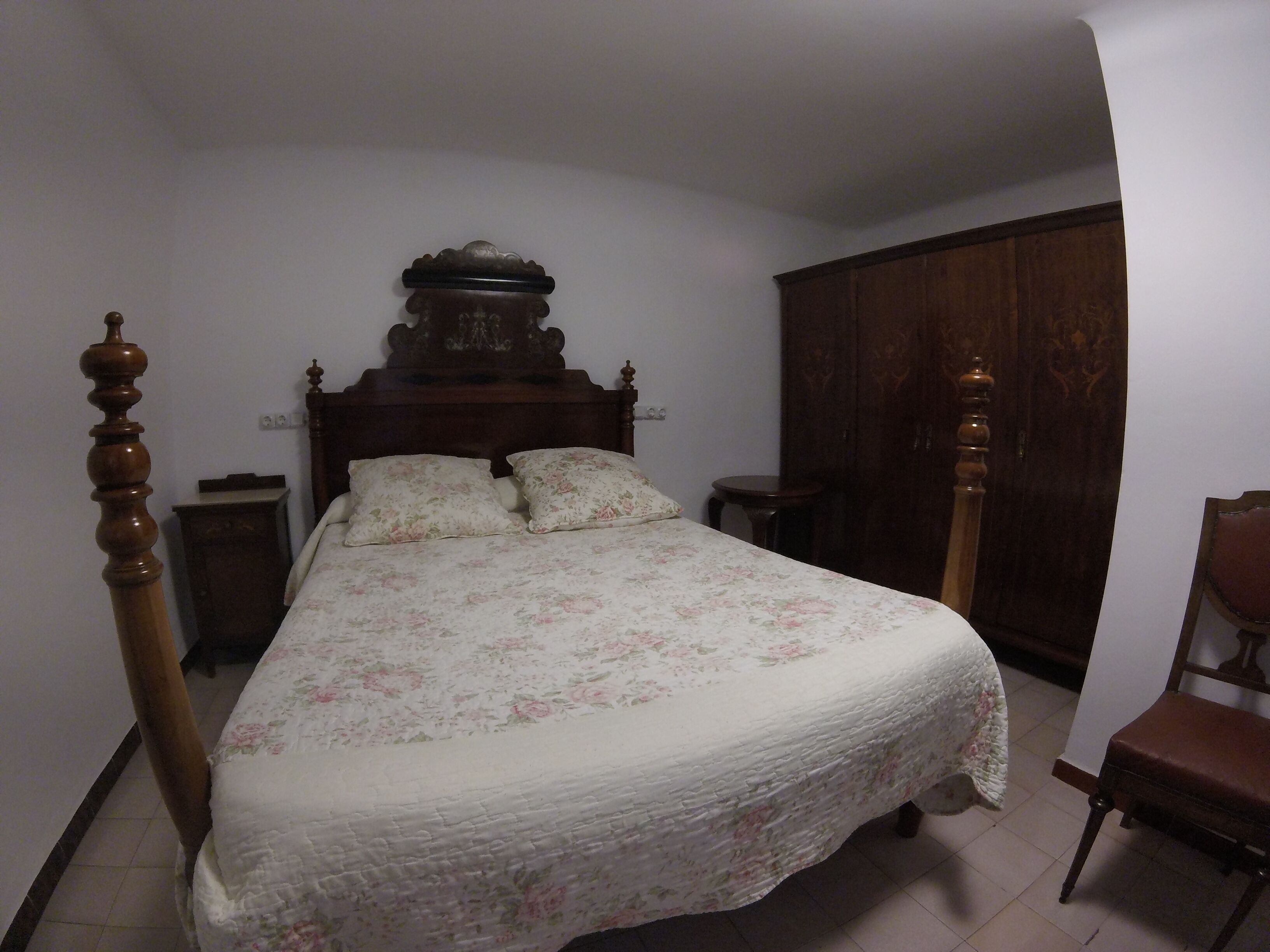 A bedroom with a wooden bed frame, floral bedding, a nightstand, a wardrobe, and a chair.