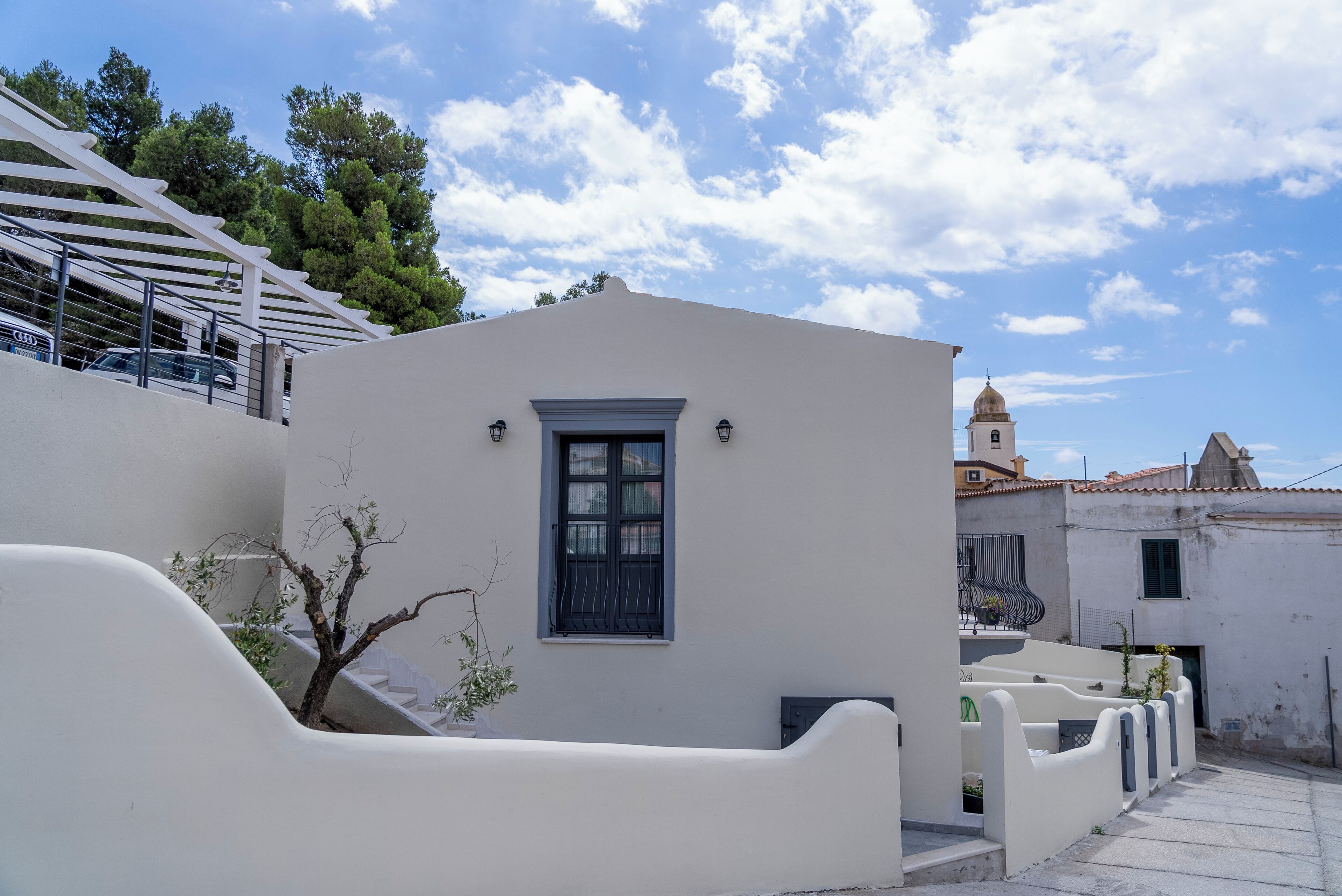 Un edificio blanco con una ventana pequeña y una puerta, rodeado por un muro blanco y un árbol.