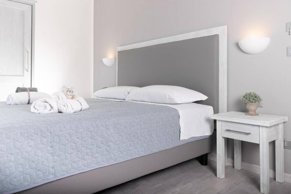 Ein ordentlich bezogenes Bett mit weißen Bettwäsche und einer grauen Decke, ein Nachttisch mit einer Pflanze sowie eine an der Wand befestigte Lampe.