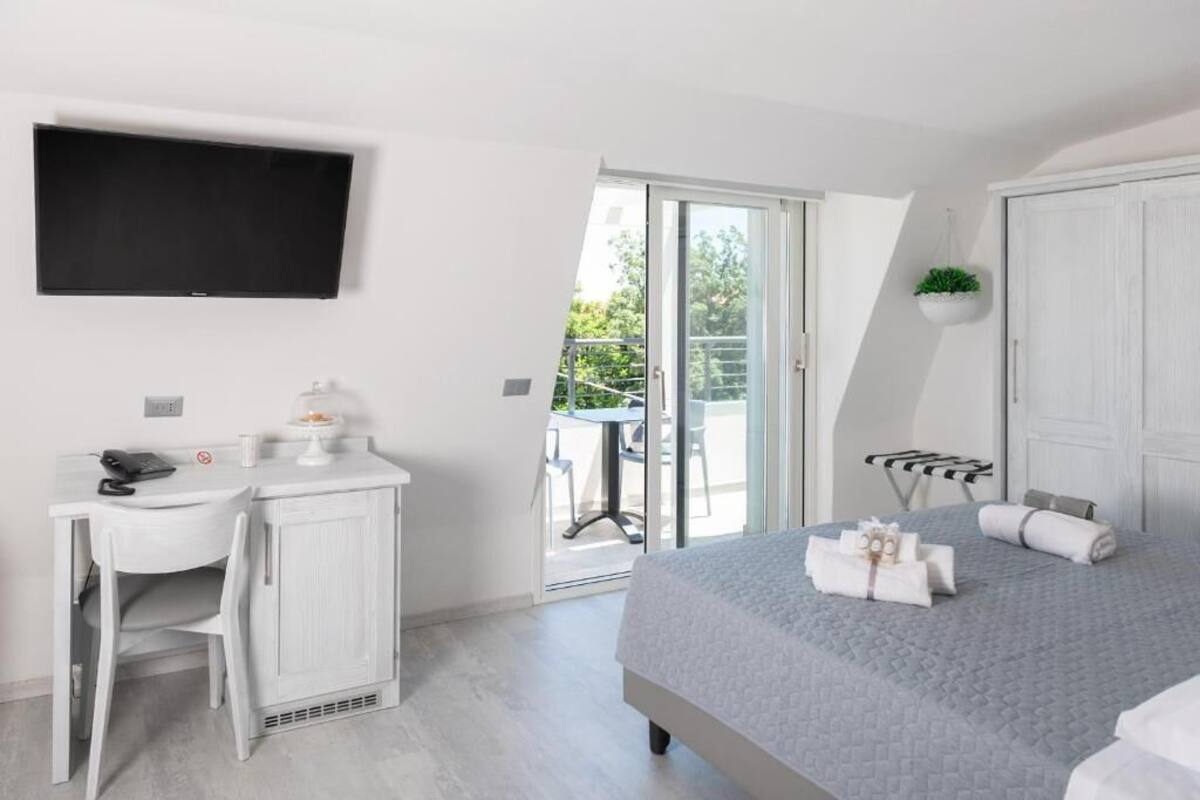 Ein modernes Hotelzimmer mit Bett, Schreibtisch, Stuhl, Fernseher und einem Balkon mit Blick auf Grünflächen.