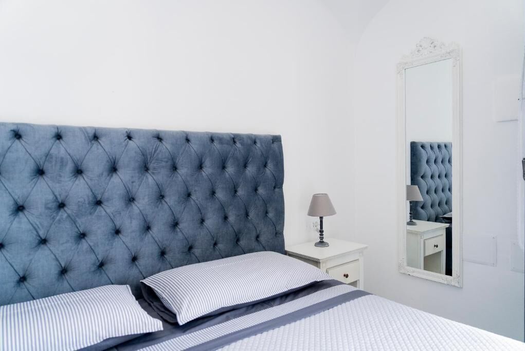 A bedroom with a tufted headboard, a bedside table with a lamp, a mirror, and a bed with striped bedding.