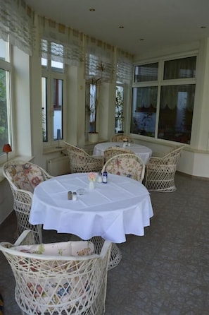 A dining area with round tables covered in white tablecloths, surrounded by wicker chairs with floral upholstery.
