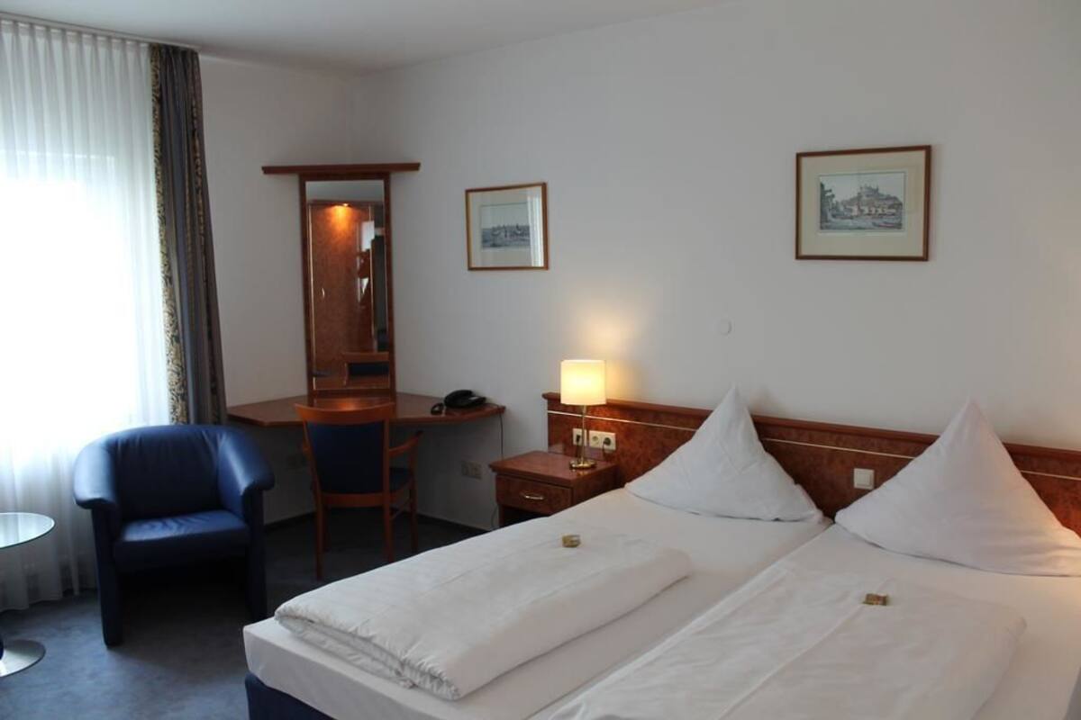 A hotel room with a bed, a desk, a chair, and two framed pictures on the wall.
