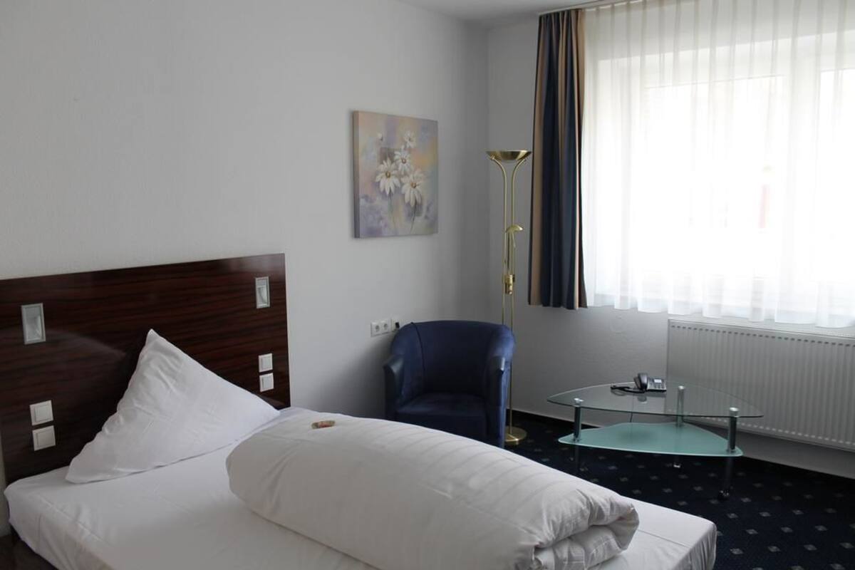 A hotel room with a bed, a chair, a small table, and a painting on the wall.