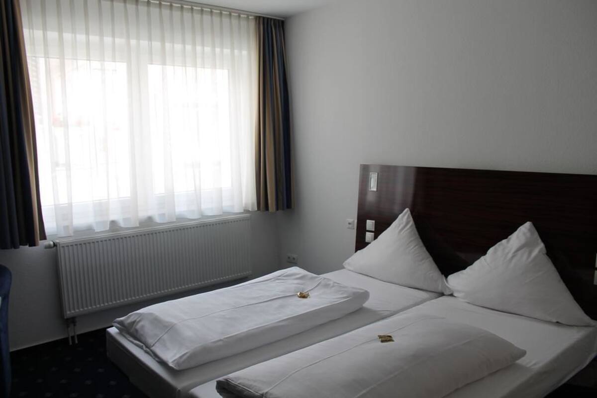 A hotel room with two single beds, a wooden headboard, a window with sheer curtains, and a radiator.