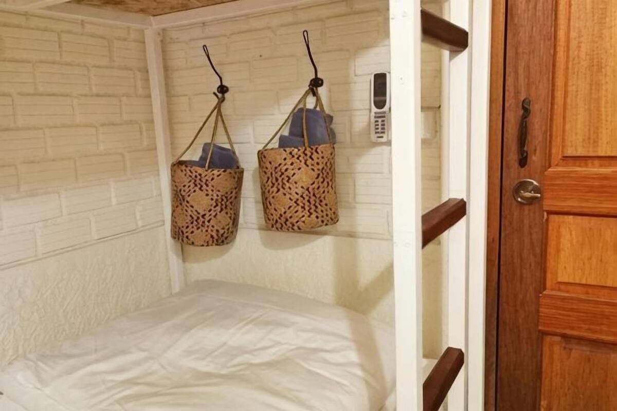 A room with a bed, two patterned bags hanging on hooks, a wooden door, and a white brick wall.