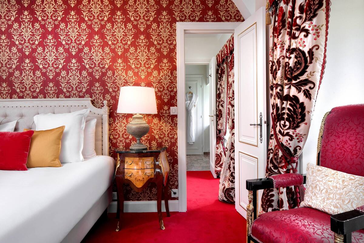 Hotelkamer met een rood tapijt, een wit bed met rode en gouden kussens, een houten nachtkastje met een lamp en een rode fauteuil.