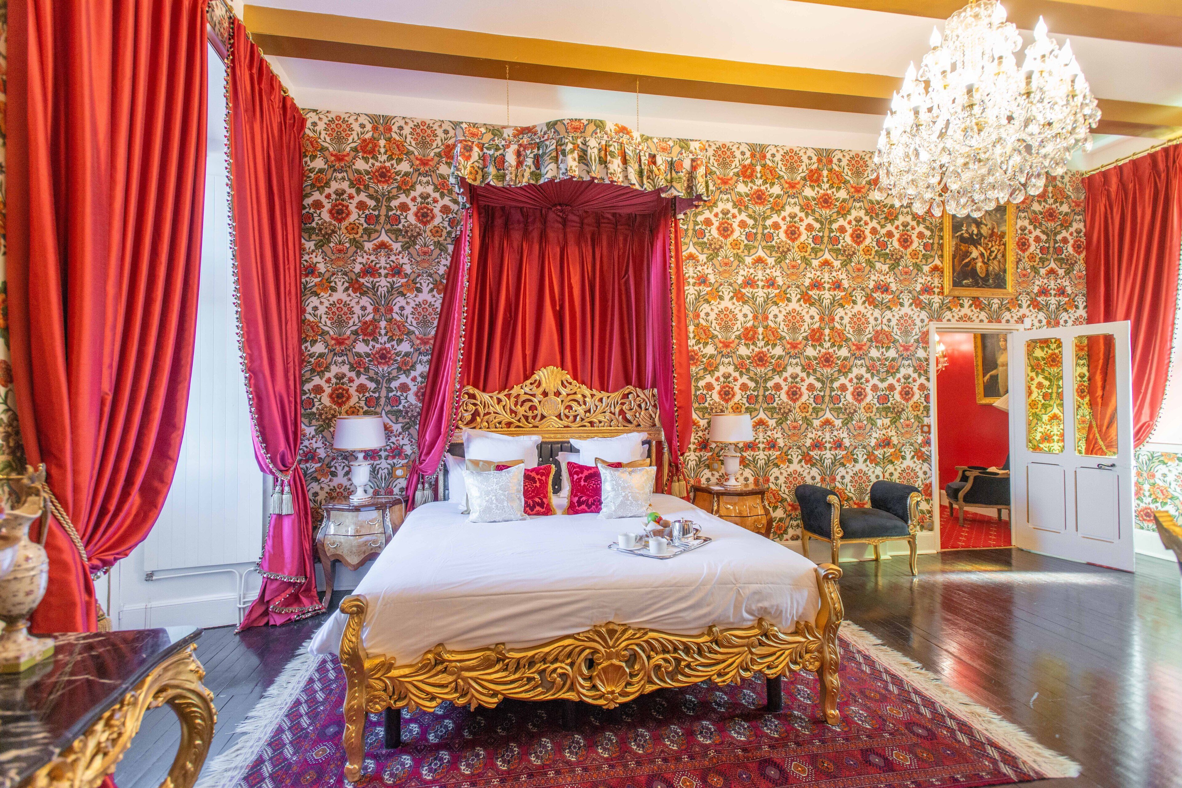 Une chambre à coucher luxueuse, avec un grand lit richement décoré, des rideaux rouges et un papier peint à motifs floraux.