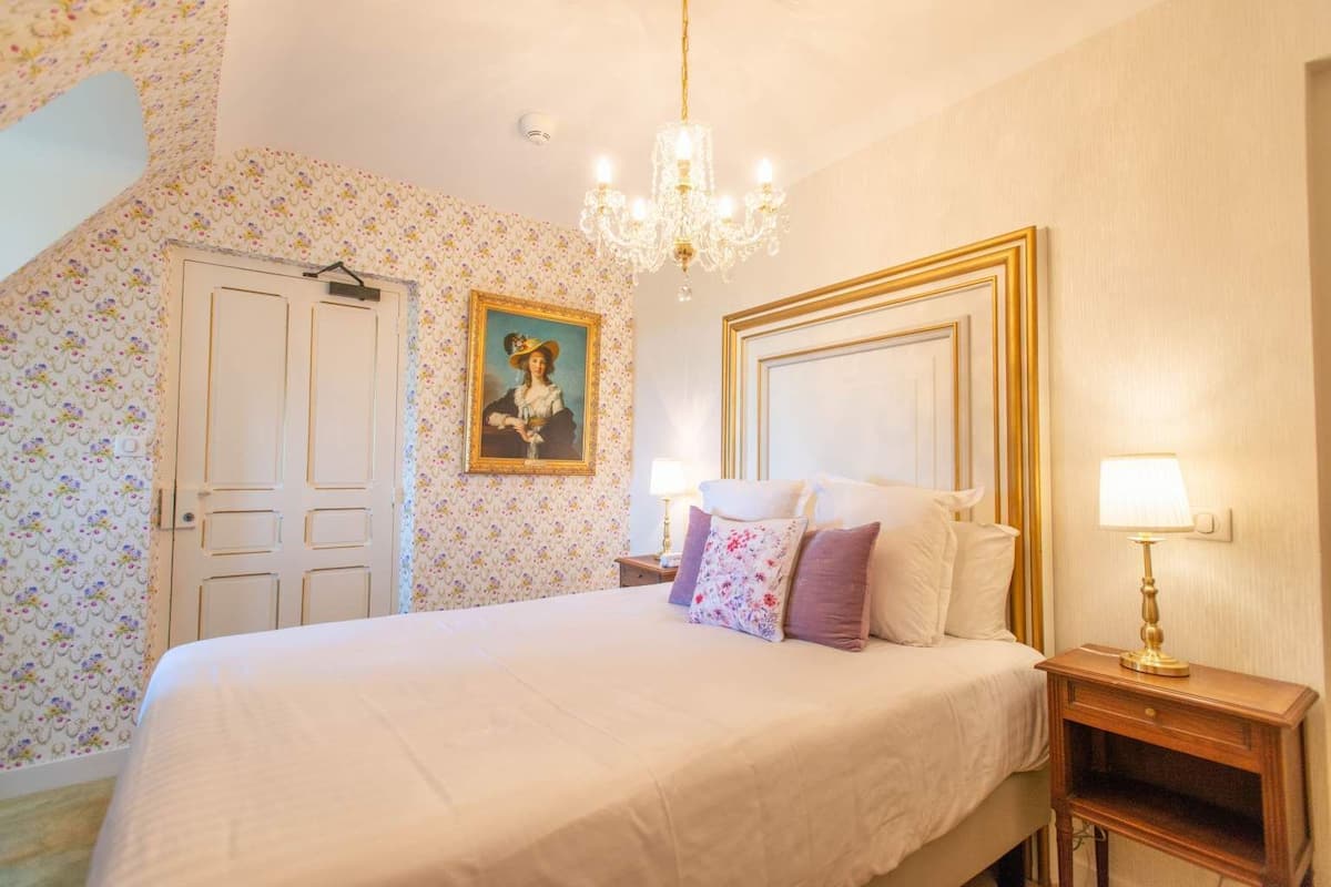 Een slaapkamer met een groot bed, een kroonluchter, een schilderij aan de muur en een deur met een bloemen behangpatroon.