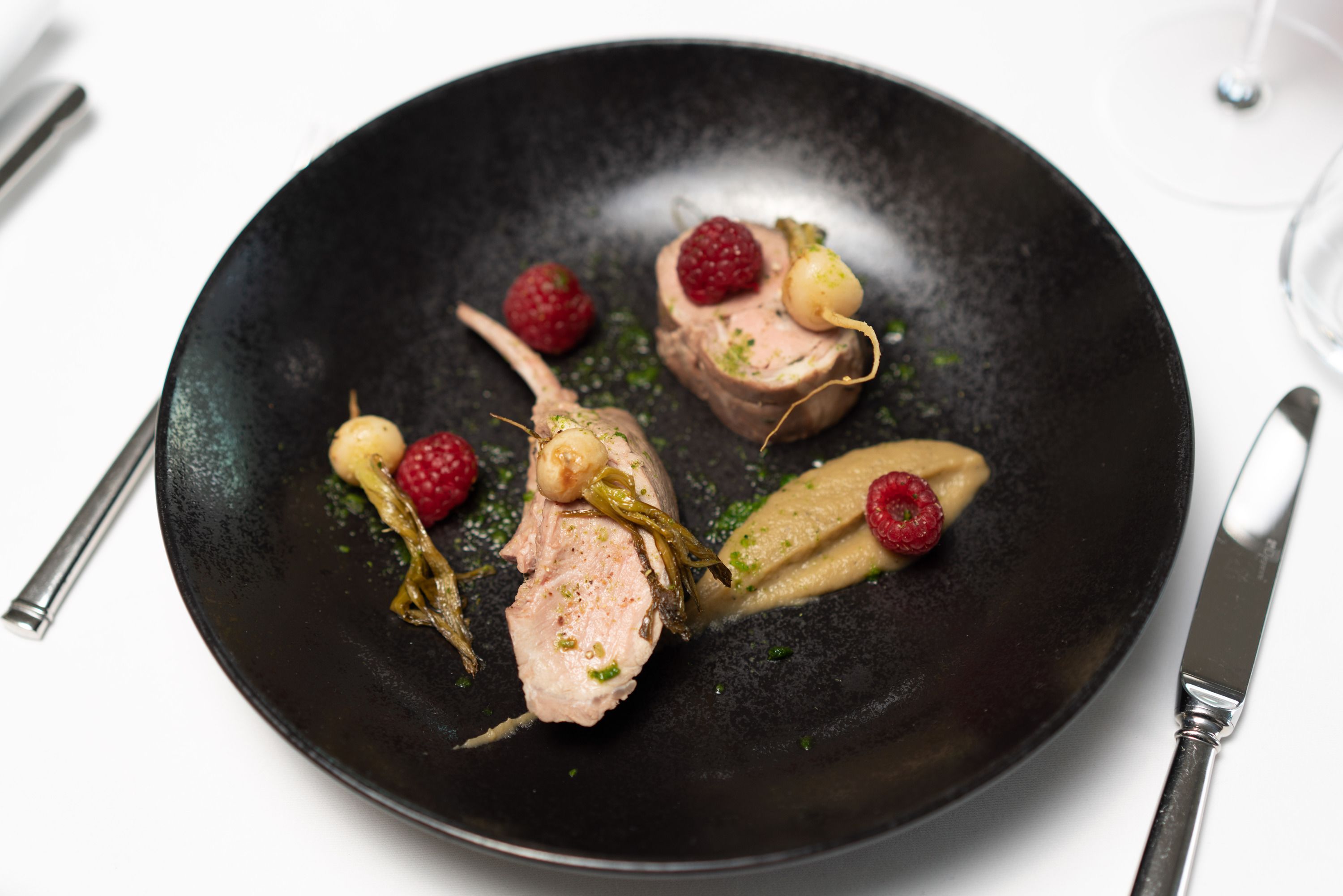 A black plate with a dish of food, including meat and berries, on a white table.
