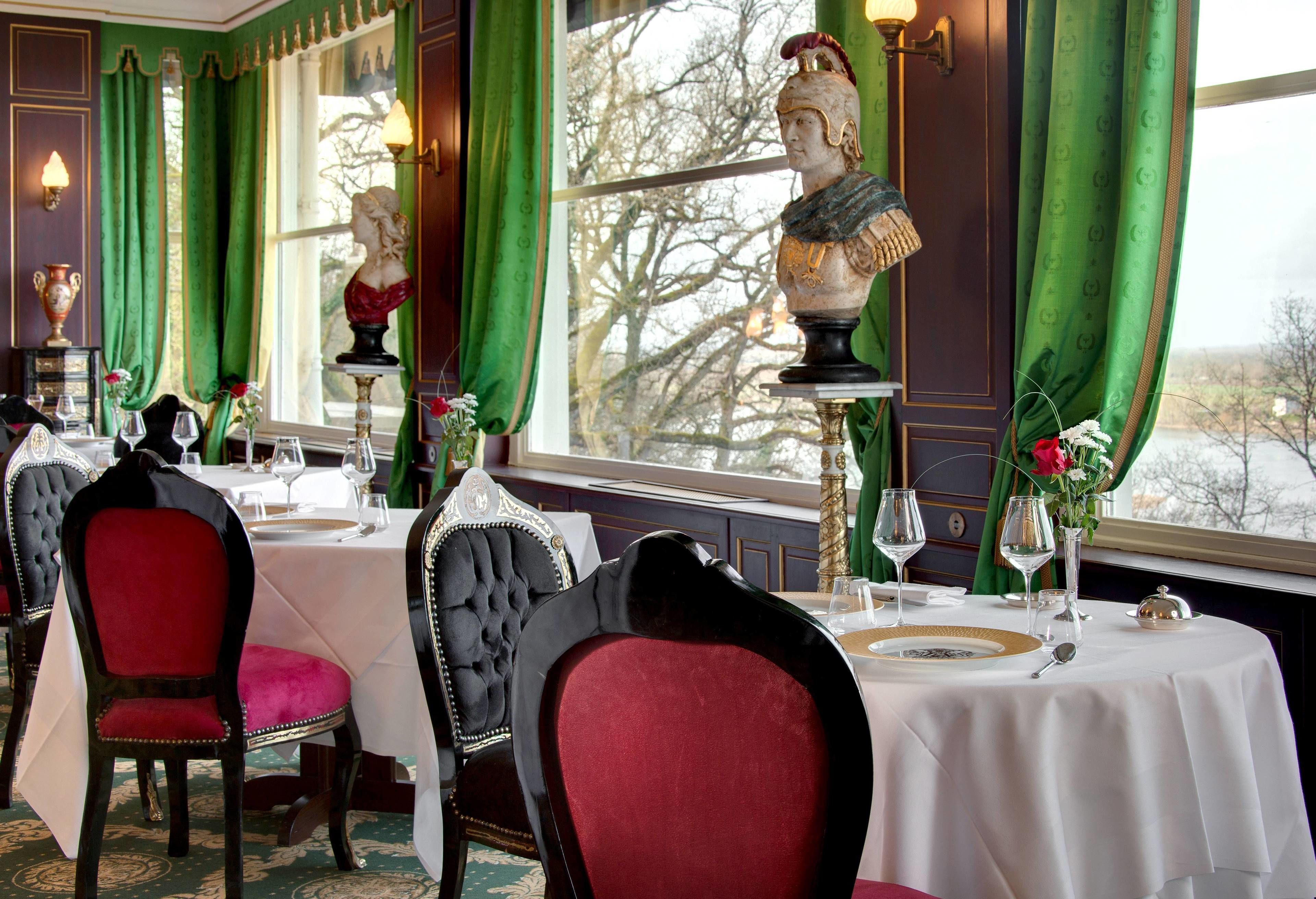 Un espace repas avec des tables préparées pour deux personnes, des rideaux verts et une statue sur un piédestal.
