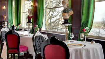 Un espace repas avec des tables préparées pour deux personnes, des rideaux verts et une statue sur un piédestal.