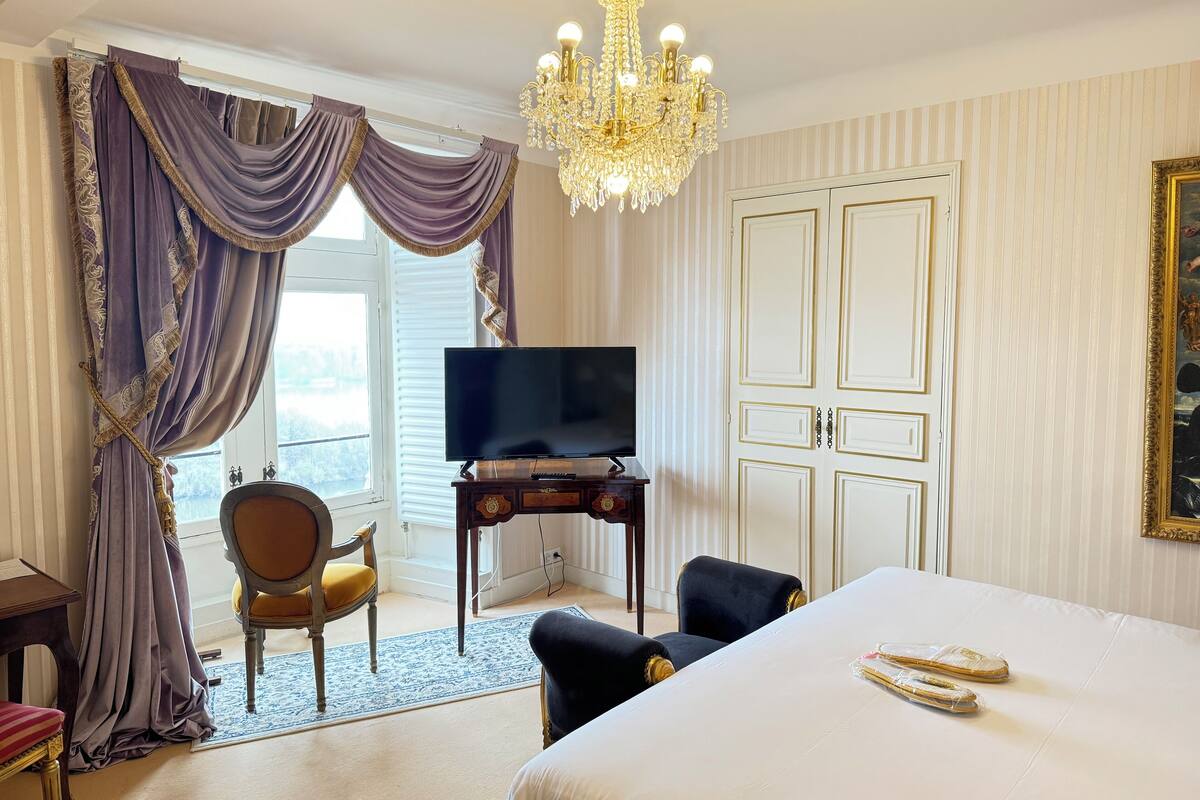 Hotelkamer met een groot bed, een televisie op een houten standaard, een kroonluchter en uitzicht op het landschap door het raam.