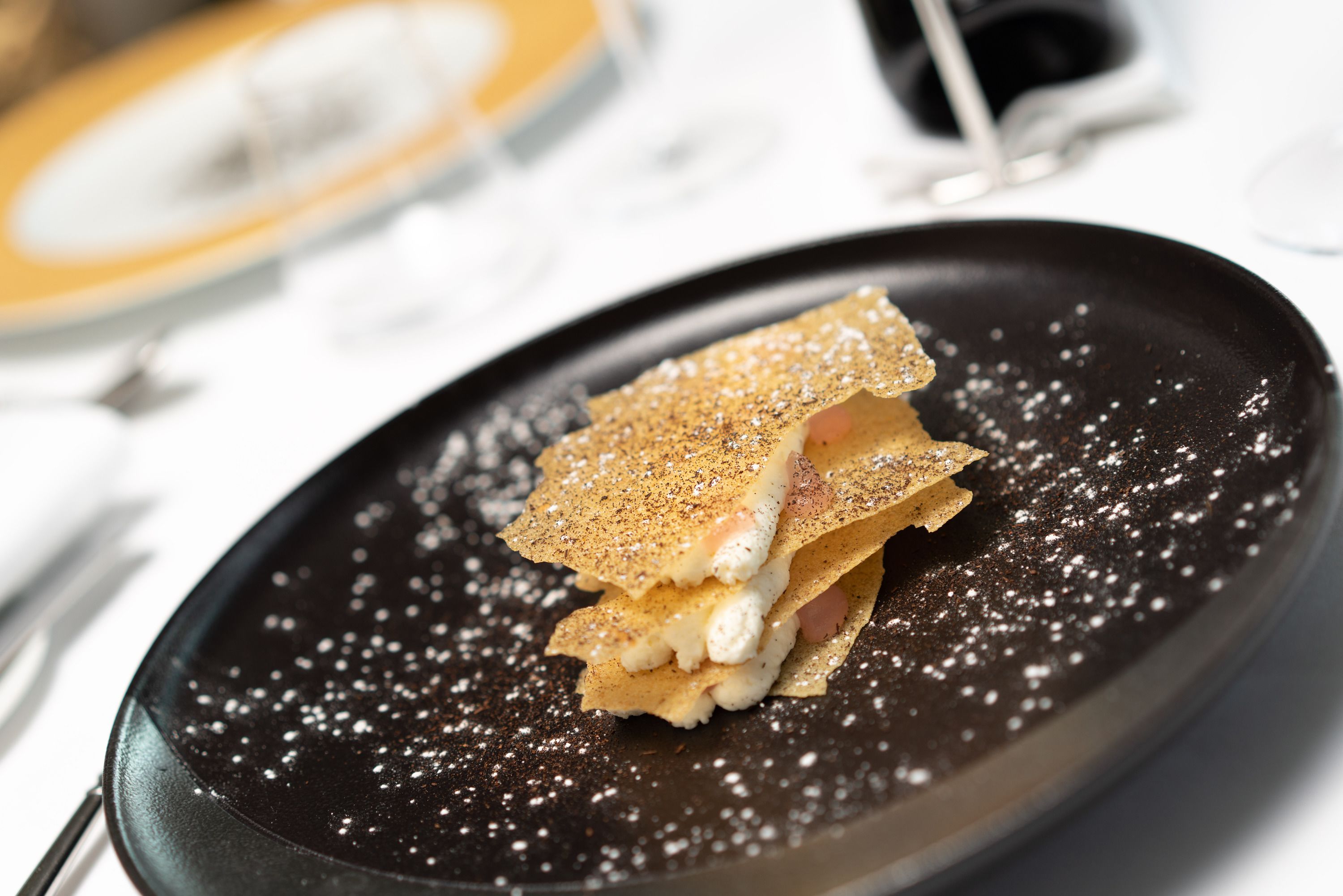 A dessert on a black plate with powdered sugar, accompanied by a fork and a knife on the table.