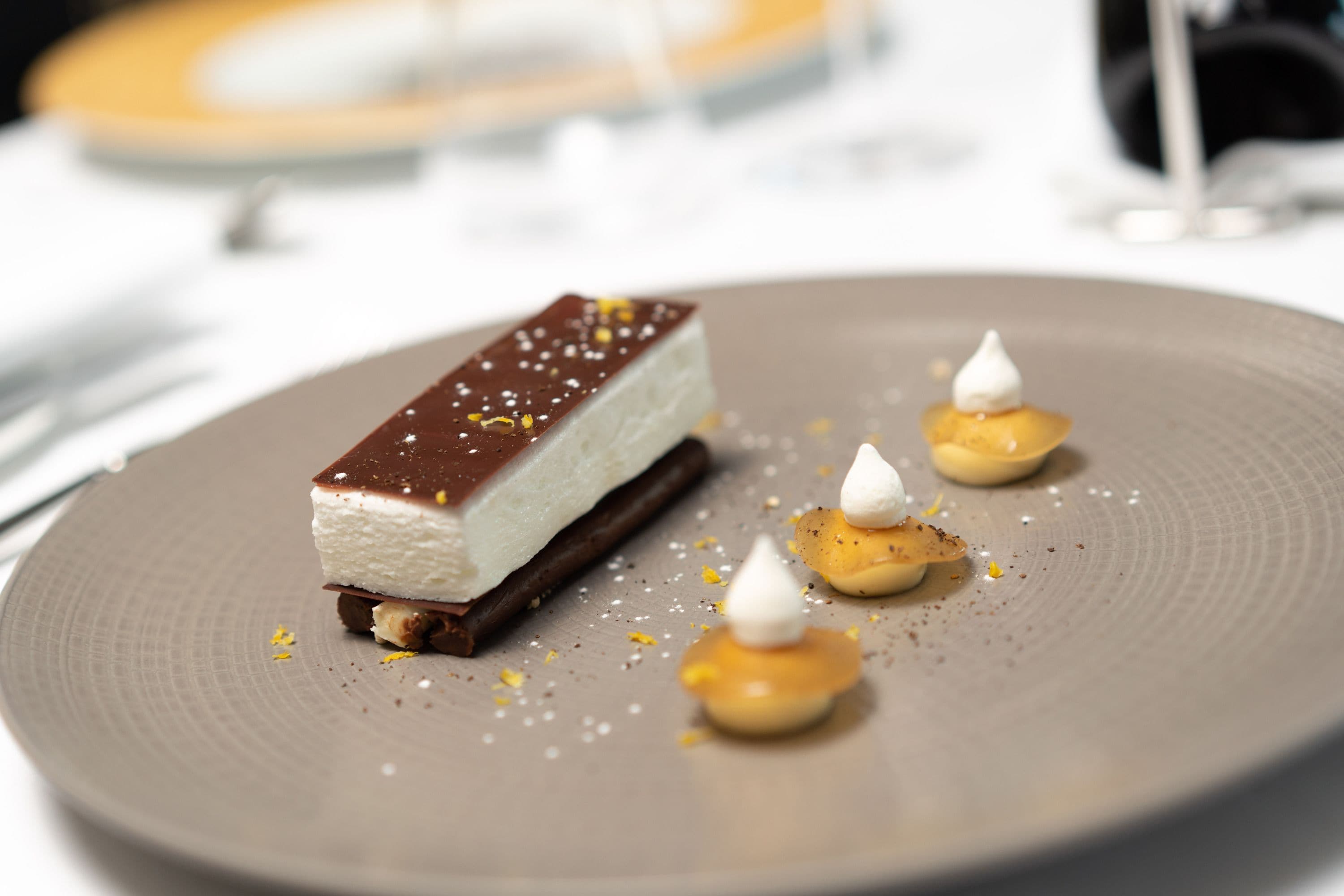 Un dessert présenté dans une assiette, composé d’une couche de chocolat, d’une crème blanche et de petites quenelles de crème surmontées d’une garniture blanche.
