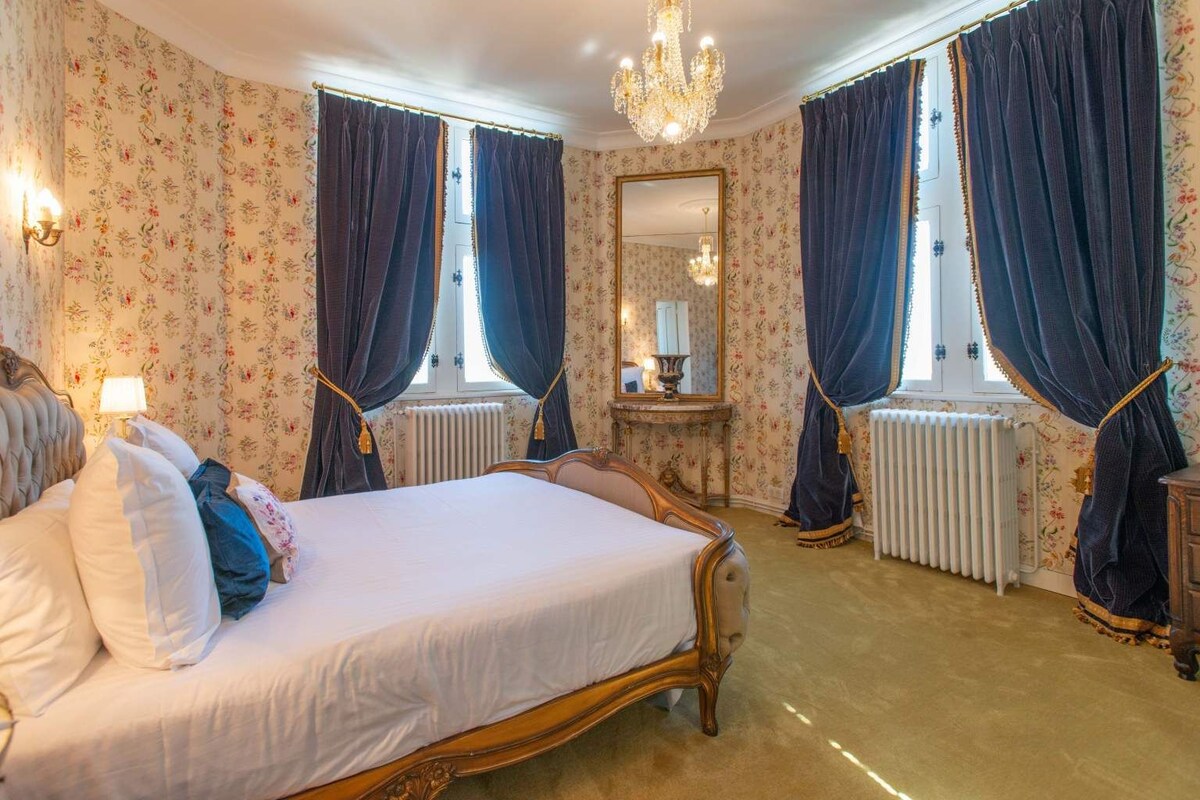 Une chambre à coucher classique, avec un grand lit, des rideaux décoratifs, un lustre et un papier peint à motifs floraux.