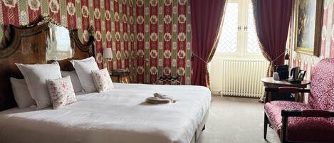 A neatly made bed with white linens and pillows, a wooden headboard, a chandelier, a red patterned wallpaper, a window with red curtains, and a red upholstered chair.