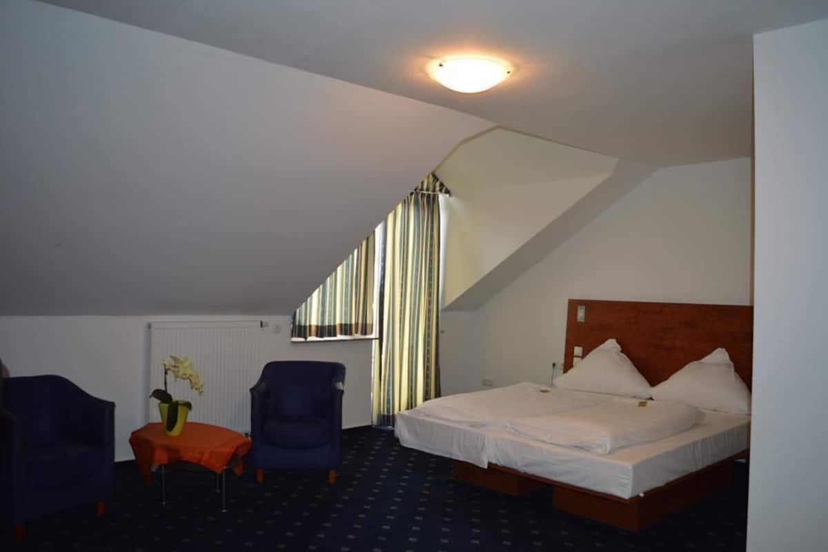 A hotel room with a bed, a chair, a small table with a vase, and a window with curtains.