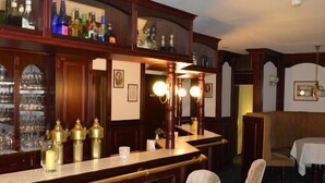 A bar area with wooden bar counters, two cushioned bar stools, and a variety of bottles and glasses on shelves. - Arosa Hotel (Kassel)