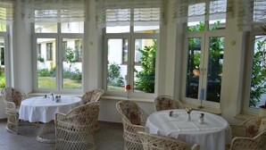 A covered outdoor dining area with wicker chairs and round tables, large windows with views of greenery, and a tiled floor. - Arosa Hotel (Kassel)