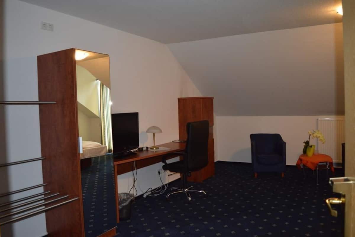 A compact hotel room with a desk, chair, and a small table with a vase.