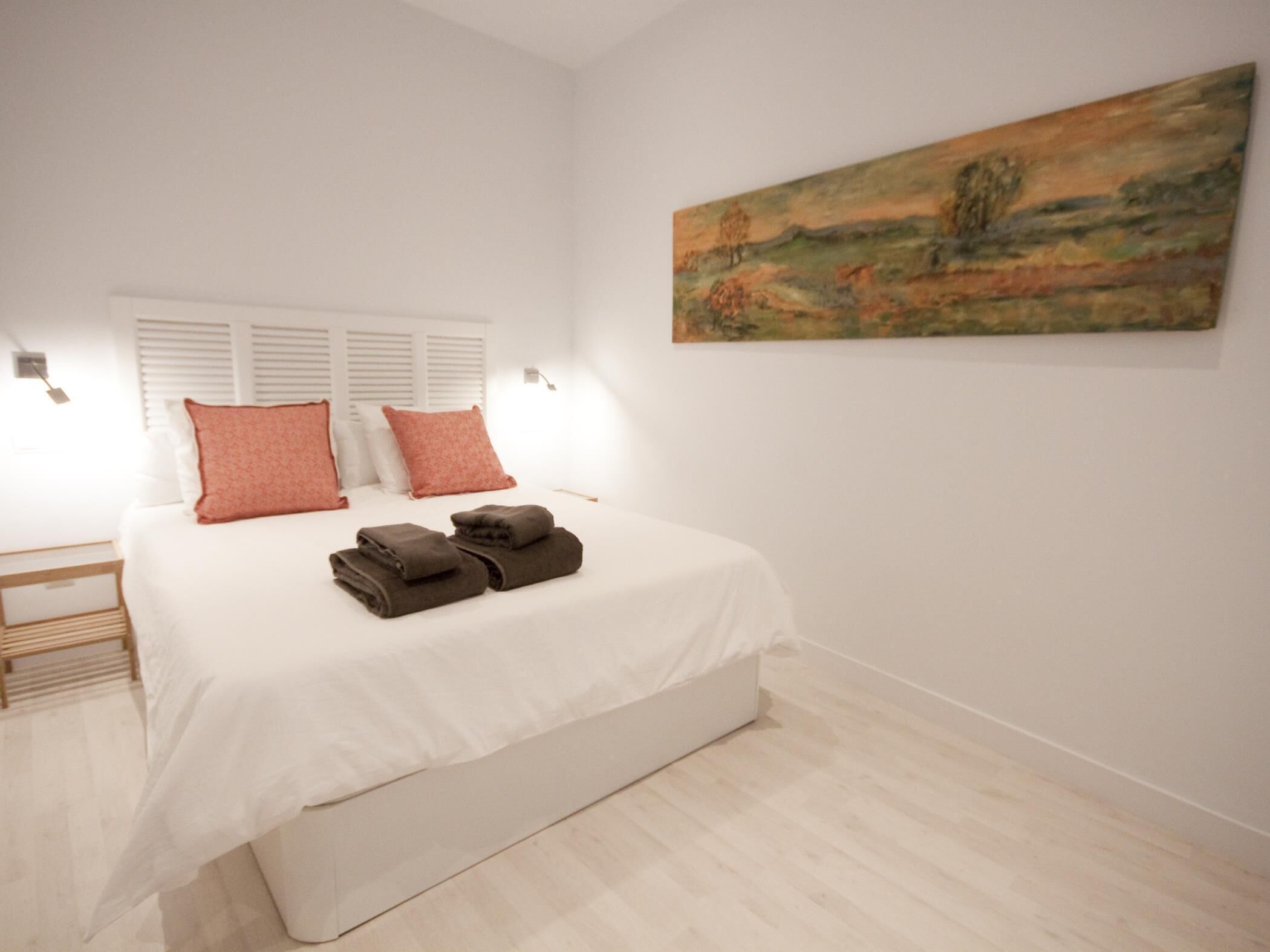 A bedroom with a large bed, white bedding, two pillows, a painting on the wall, and a bedside table with a lamp.