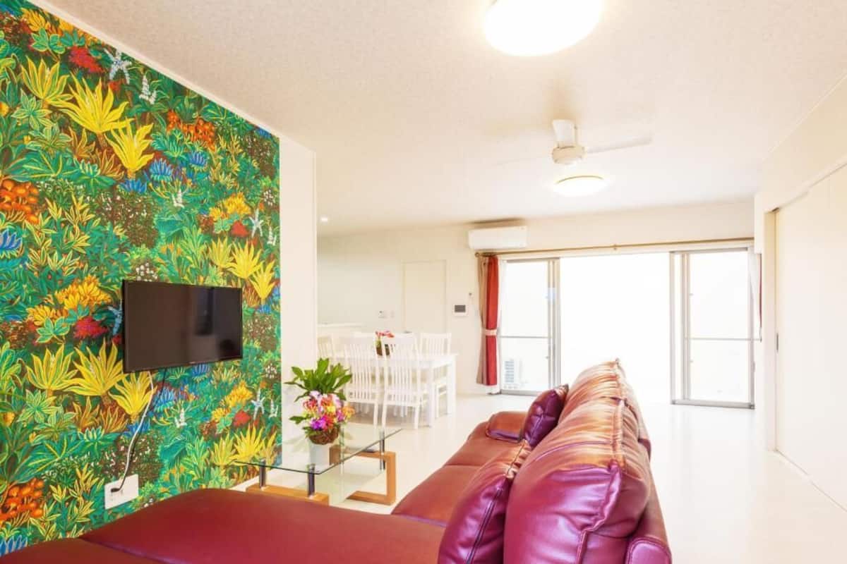 Een woonkamer met een rode bank, een televisie bevestigd aan een kleurrijke muur, en een eethoek met witte meubels.