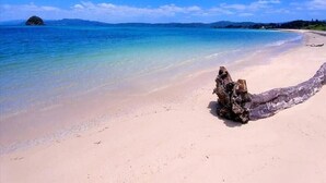 Une plage à l’eau turquoise et limpide, une grande formation rocheuse et, au loin, des montagnes sous un ciel bleu.