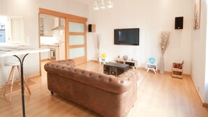 Una sala moderna con un sofá marrón, una mesa de comedor, una cocina al fondo y un televisor de pantalla plana montado en la pared.